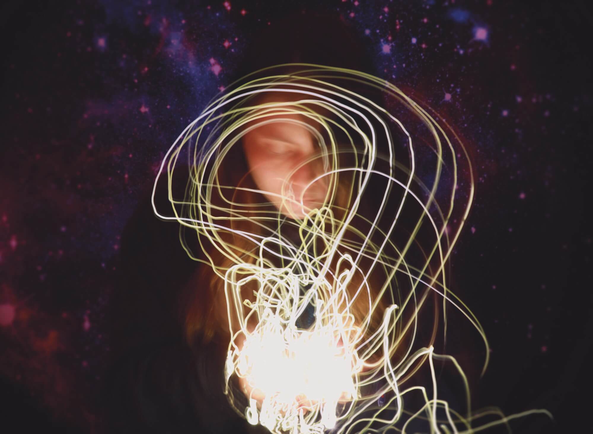 A person surrounded by swirling trails of light against a starry sky backdrop. The lights are predominantly white and yellow, creating dynamic, looping patterns around the individual.